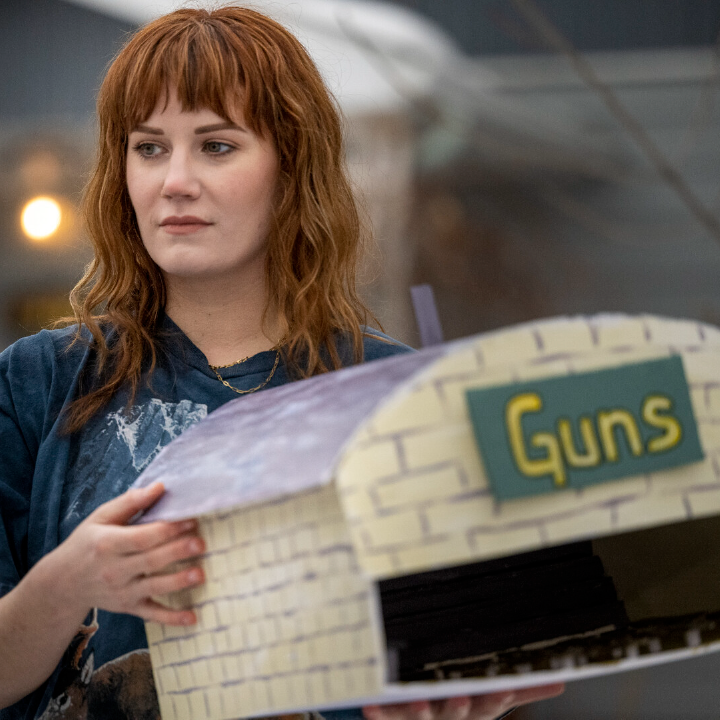 Person holding a model of a building that says "GUNS" on the front